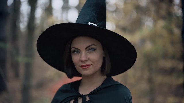 Beautiful witch in cap, long dress, mantle on autumn forest background. Halloween concept, cosplay dressing up. Slow motion