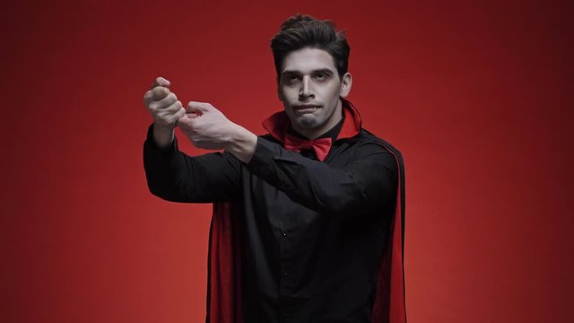 Vampire man with blood and fangs in black halloween costume gesturing like he is trying to kill himself with a gun and then stab with a knife isolated over red wall