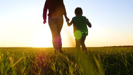 A young mother holds her son's hand and runs through the green grass against a beautiful sunset.