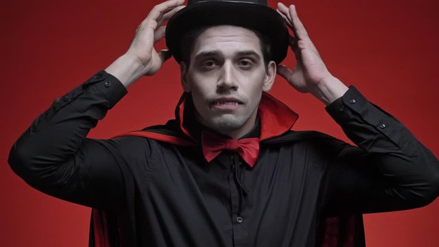 Attractive vampire man with blood and fangs in black halloween costume putting on his hat, fixing his bow tie and cloak isolated over red wall