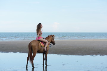 A pretty woman in a bikini is happily riding a horse on the beach.