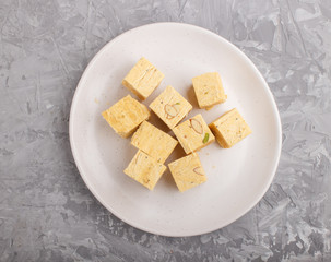 traditional indian candy soan papdi in white plate with almond and pistache on a gray concrete background. top view