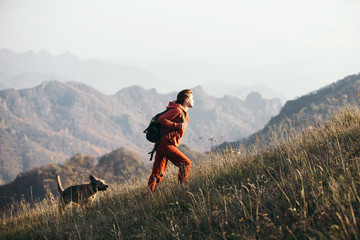 Beautiful woman traveler climbs uphill with a dog on a background of mountain views. She is with a backpack and in red clothes.