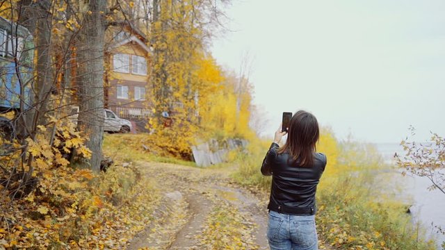 A girl in a leather jacket on the banks of the river takes pictures of nature