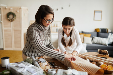 Young woman in eyeglasses decorating Christmas present with wrapping paper together with little...