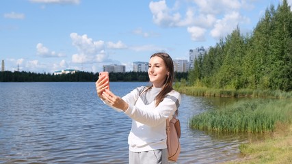 Beautiful girl with a backpack takes a selfie near the lake and talks on a cell phone on Skype.