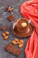 Cup of hot chocolate and pieces of milk chocolate with almonds on a black concrete background with red textile. side view.