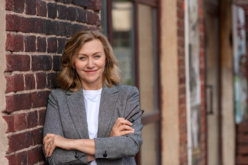 Portrait of a beautiful blonde woman, 55 years old, smiling. A business woman is standing against a red brick wall at the entrance to her store.