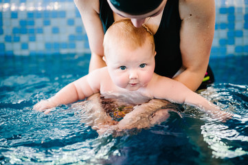 Fototapeta na wymiar Young mother, swimming instructor and happy little girl in the pool. Teaches infant child to swim. Enjoy the first day of swimming in the water. Mom holds child preparing for diving. doing exercises