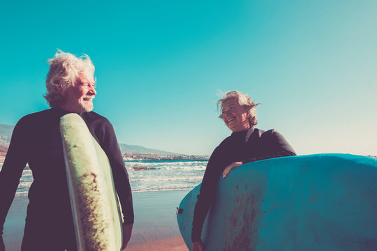 couple of seniors at the beach with black wetsuits holding a surftable ready to go surfing a the beach - active mature and retired people doing happy activity together in their vacations or freetime
