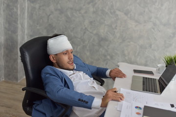 Overworked businessman with bandaged hurt head at workplace with scattered documents
