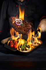 Chef cooking steak and vegetables in pan with fire flame on  dark background. Restaurant and hotel...
