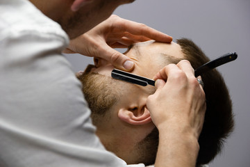 Barber shaving client beard with sharp razor blade in close up.
