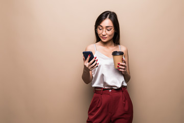 Full length portrait of a smiling asian woman using mobile phone while holding cup of coffee to go isolated over beige background