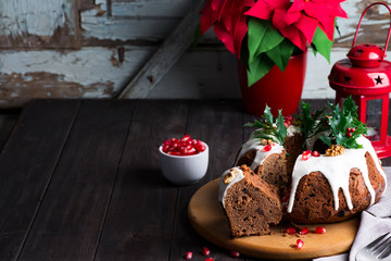 Christmas sliced chocolate cake with white icing, holly branches and pomegranate kernels on a wooden dark background