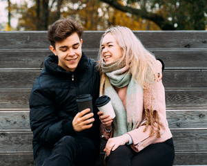Lovely brunette guy and pretty girl blonde drink coffee and chat on date, laugh. Loving teenagers are sitting on park bench. Teen Love Concept. They are happy, smiling, laughing