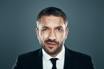 Charming young man in full suit looking at camera while standing against grey background