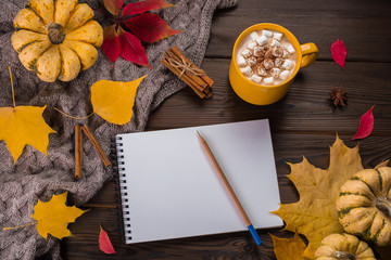 Naklejka na ściany i meble Autumn flat lay. Pumpkins and yellow leaves on knitted plaid. Wooden background. Yellow cup with coffee and marshmallow. White paper notepad with pencil.