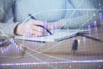 Hands writing information about stock market in notepad. Forex chart holograms in front. Concept of...