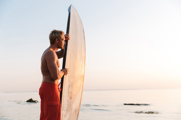 Image of blonde surfer man holding his surfboard by ocean at sunrise