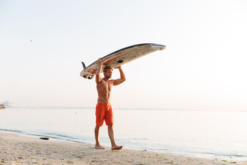 Image of muscular surfer man carrying his surfboard by ocean at sunrise