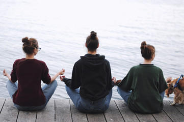  Three young women doing yoga at nature. Fitness, sport, yoga and healthy lifestyle concept - group of people making yoga pose on lake pier at sunset
