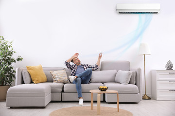 Young man with remote control suffering from heat under broken air conditioner at home