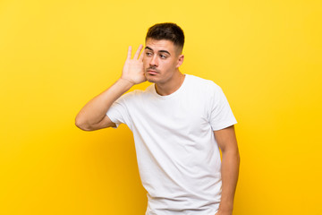 Fototapeta na wymiar Young handsome man over isolated yellow background listening to something by putting hand on the ear