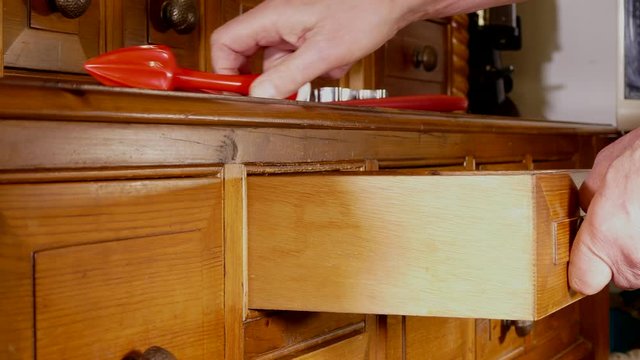A close POV dolly shot of a man&rsquo;s hands putting away kitchen utensils into the wooden drawers of a traditional, old pine dresser. 