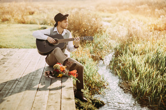 Young man playing guitar sitting on the bank of a mountain river on a background of rocks and forest. Handsome hippie style guitarist engrossed on music outdoors. Concept of freedom relaxation. Place