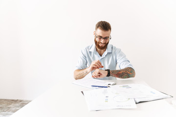 Image of young man looking at wristwatch while working at table in office