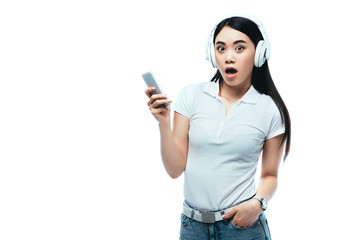 shocked attractive asian girl in headphones using smartphone isolated on white