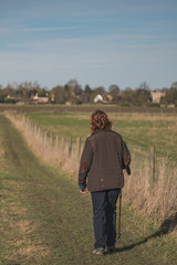 Adult Woman seen walking with the aid of a Walking Pole, in a rural location in the East of England. She is walking adjacent to a nature reserve, the distance shows a rural community and village.