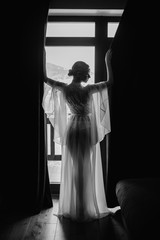 Black and white photo of bride in peignoir standing by the window. Bride morning