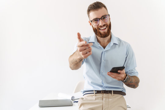 Image of optimistic young man holding smartphone while working in office