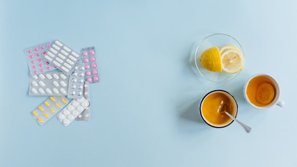 On the right side cup of tea, saucer of lemon and honey. On the left side assorted pharmaceutical medicine multicolored pills. Choice of treatment method. Healthcare concept. Flat lay, copy space.
