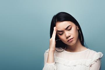 upset brunette asian woman with headache on blue background