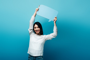 happy brunette asian woman holding speech bubble on blue background