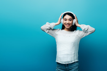 smiling asian woman with hands on headphones on blue background