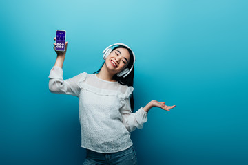 smiling asian woman with closed eyes in headphones holding smartphone with healthcare app and dancing on blue background