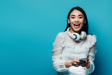 happy asian woman with wireless headphones in white blouse using smartphone on blue background