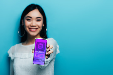 selective focus of smiling asian woman in white blouse holding smartphone with online shopping app on blue background