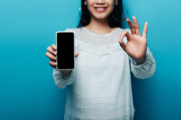 cropped view of smiling asian woman in white blouse showing ok sign and smartphone with blank screen on blue background