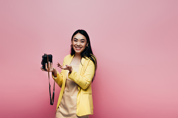 happy asian woman in yellow outfit holding digital camera isolated on pink