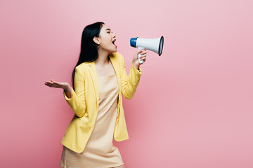 side view of asian woman in yellow outfit screaming in megaphone isolated on pink