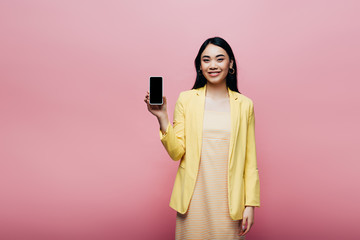 happy asian woman in yellow outfit holding smartphone with blank screen isolated on pink