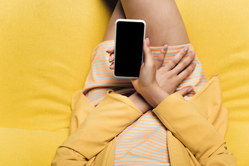 overhead view of businesswoman sitting on yellow sofa and holding smartphone with blank screen