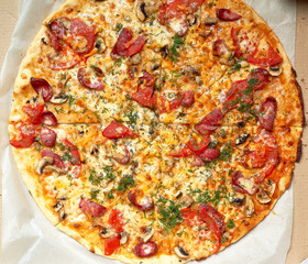 baked round pizza with smoked sausages, mushrooms, tomatoes, cheese and dill in an open cardboard box
