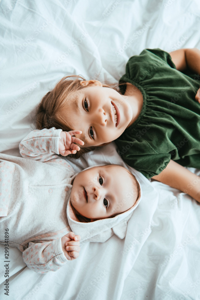 Wall mural top view of smiling child looking at camera while lying on white bedding near little sister