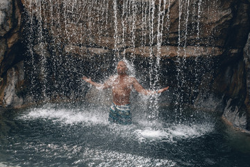 Fototapeta na wymiar Fit and handsome topless Arabic man under the waterfall, huge rocks and water, splashes and flecks; natural beauty concept.
